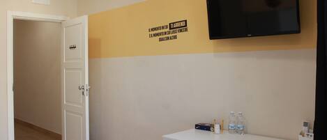 A hotel room with a white desk, a flat-screen TV mounted on the wall, a door, and a wall with a motivational quote.