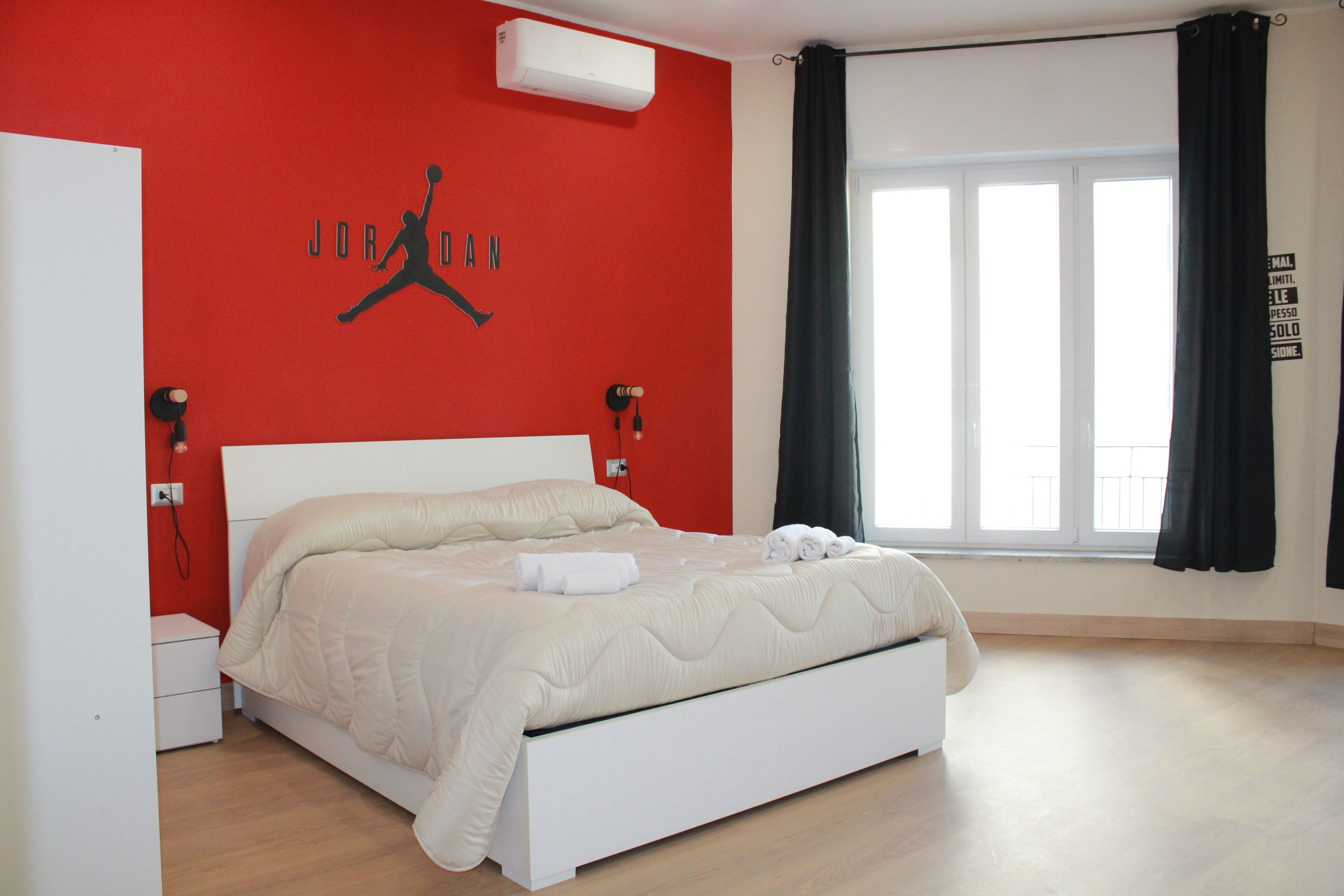 A bedroom with a bed, a red wall with a Jordan logo, a window with curtains, and a white bedside table.