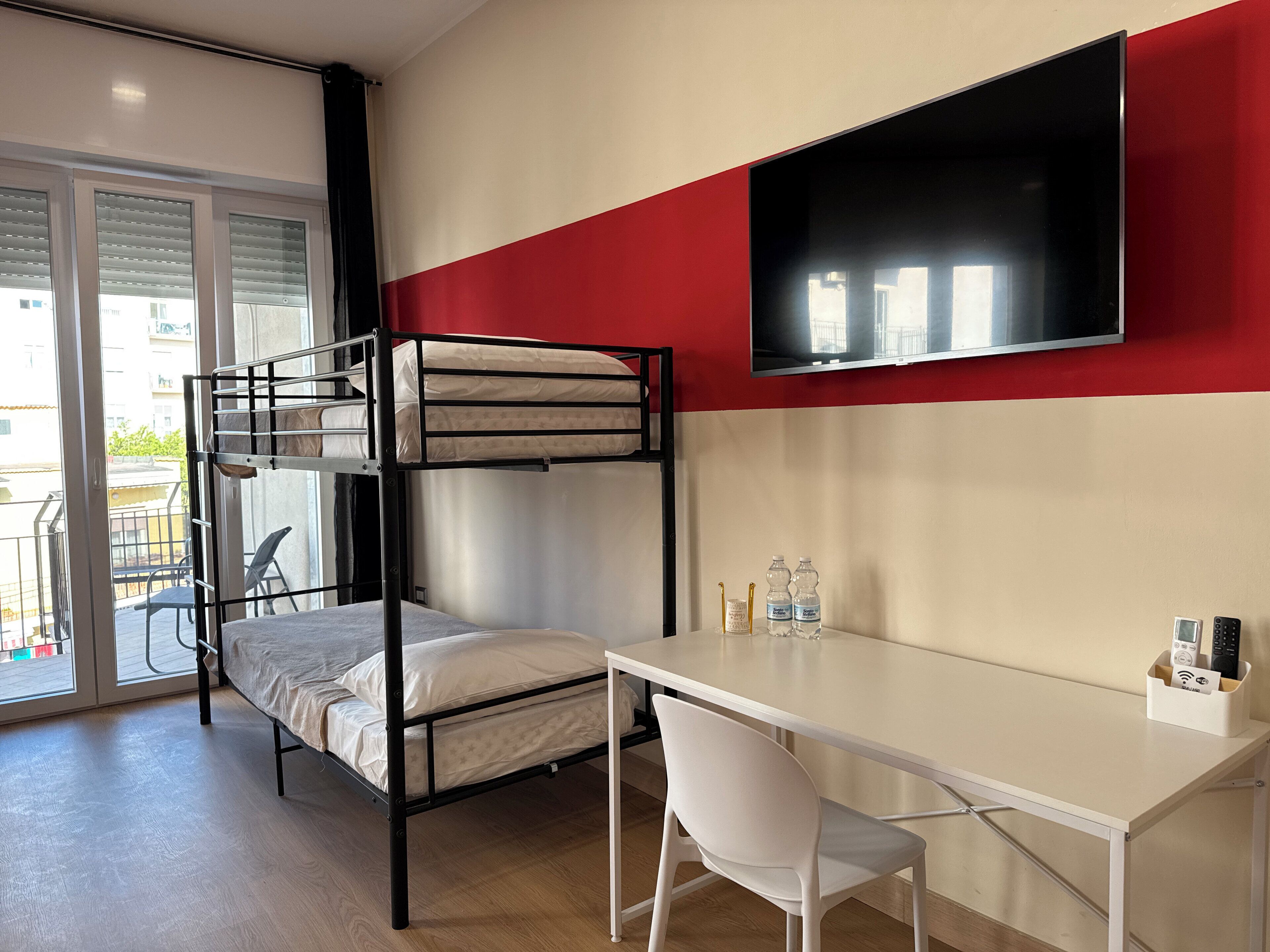 Una cama litera con escritorio y silla, un televisor en la pared y una ventana con persianas.