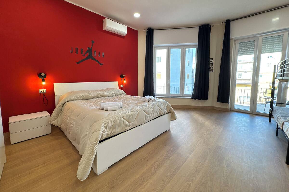 Un dormitorio con una pared roja, una cama blanca y una ventana con vistas a edificios.