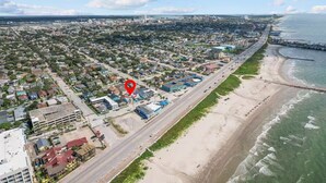 An aerial view of a coastal city with a beach, a marked location, and a road running parallel to the shore. - Coral Bungalow (Galveston)