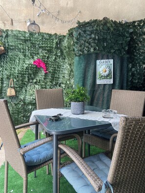 Une terrasse de jardin avec des meubles en osier, une table en verre et une pancarte sur laquelle on peut lire : « C’EST ICI QUE L’ESPOIR ET LE RENOUVEAUEMENT JARDINIERS SE TROUVENT ».