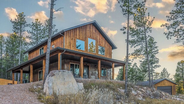 Eine zweistöckige Holzhütte mit großen Fenstern und einer Veranda, umgeben von Bäumen und einer felsigen Landschaft.