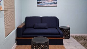 Een woonkamer met een donkerblauwe bank, een bevestigde televisie, een airconditioner, een schilderij van een zonsondergang en twee ronde bijzettafels.
