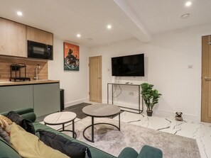A modern living room with a green sofa, two round tables, a flat-screen TV, a kitchen area with a microwave, and a wooden door.