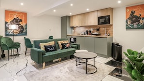 A modern living room with a green sofa, a round coffee table, and a kitchen area with wooden cabinets and a microwave. - 2A Ruby Street (London)