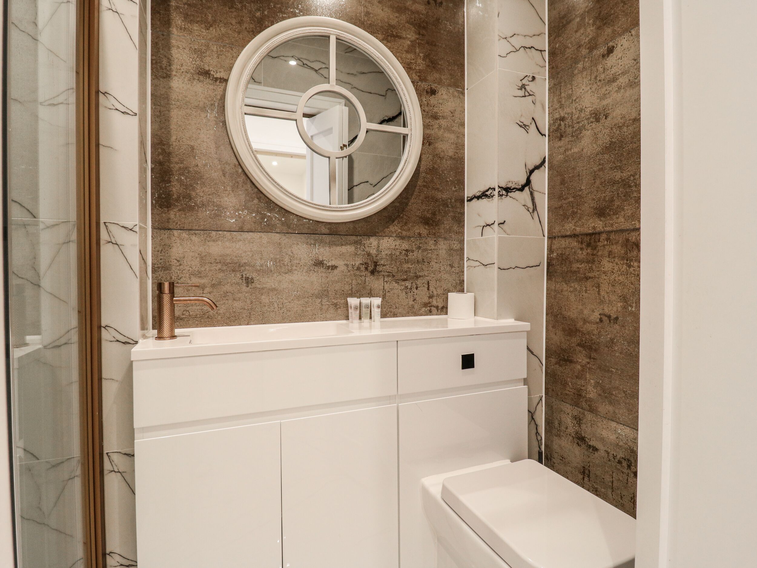 A bathroom with a marble-tiled wall, a round mirror, a white sink, and a toilet.
