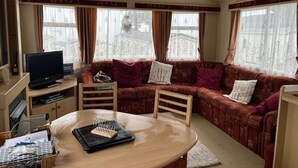 A cozy living room with a sofa, a television, a table with a notebook, and a window with curtains. - 2bed-cabin Sleeps 4, Wifi, Free Parking, Sea Views (Bembridge)