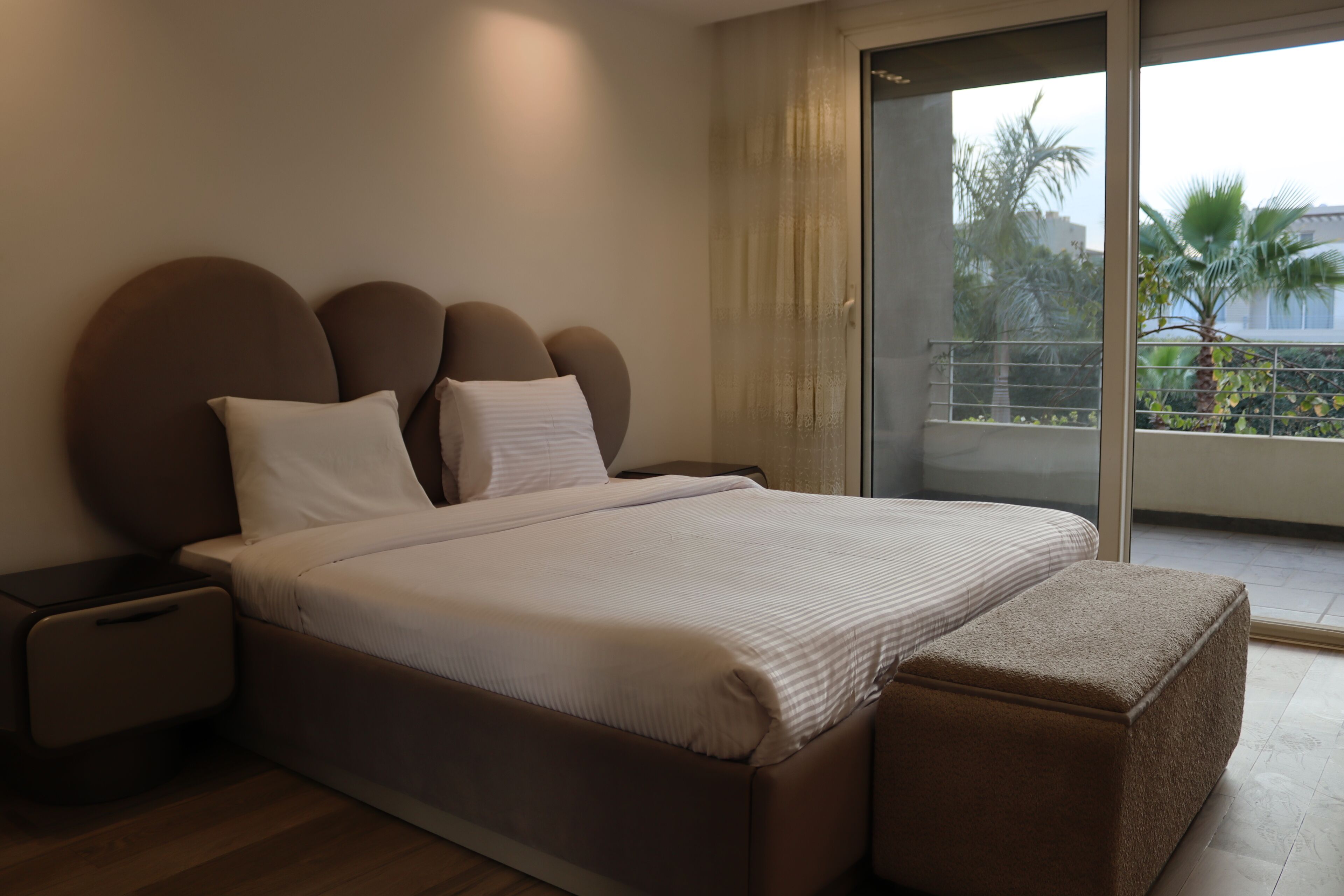 A modern bedroom with a large bed, a nightstand, and a view of a balcony with palm trees.