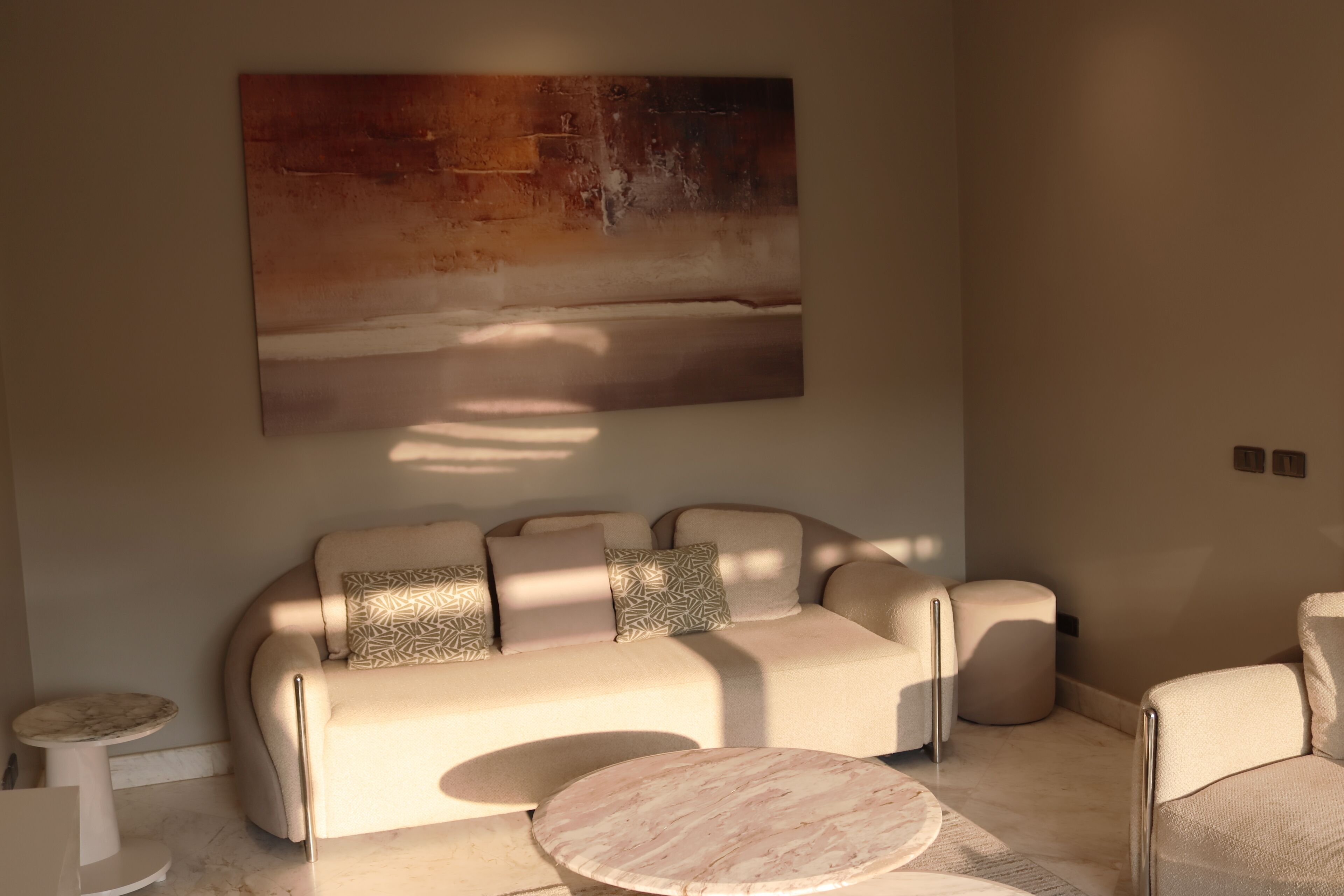 A modern living room with a beige sofa, patterned cushions, a marble coffee table, and a large abstract painting on the wall.