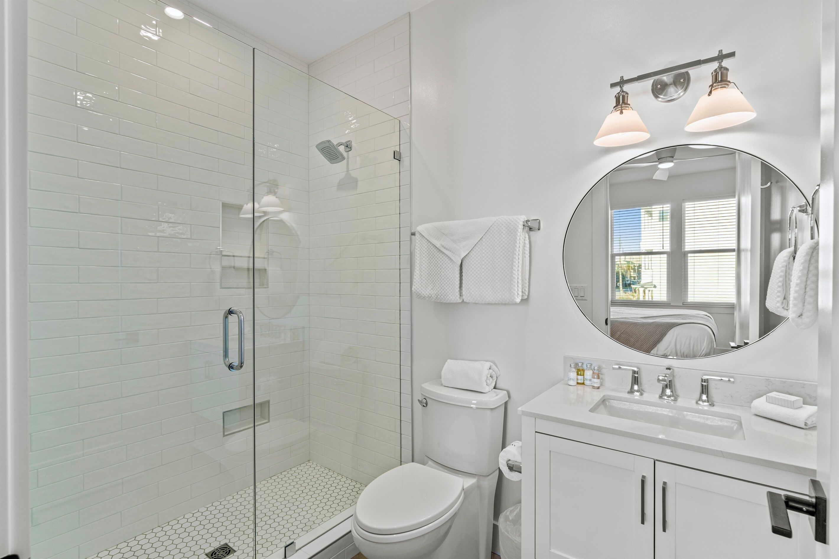 A modern bathroom with a glass-enclosed shower, a white toilet, a round mirror, and a white sink with two lights above it.