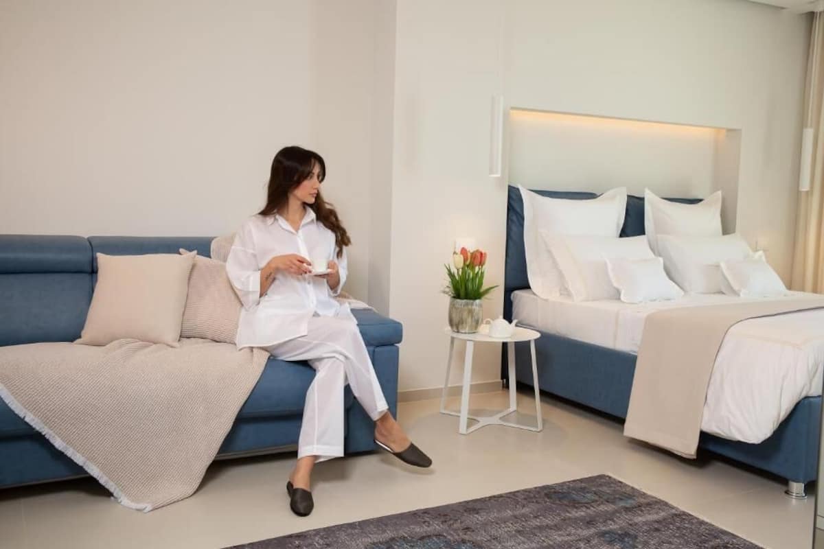 A modern hotel room with a blue sofa, a bed with white bedding, a small side table with a plant, and a coffee cup.
