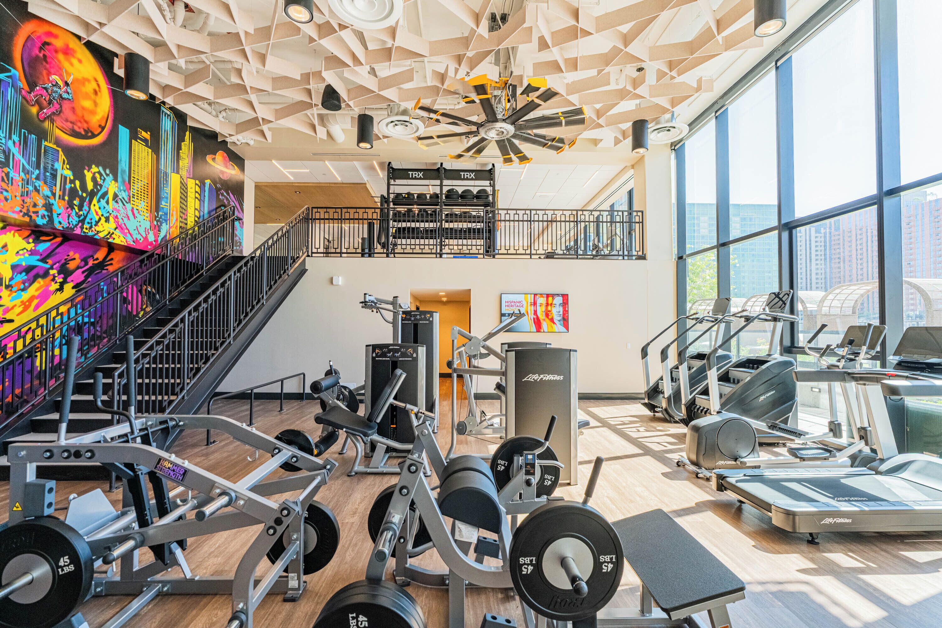 A gym with various exercise equipment, a staircase, and a large mural on the wall.