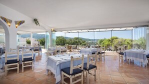 An outdoor dining area with tables and chairs, a view of the landscape, and a covered section with a glass ceiling. - Hotel with swimming pool near Baja Sardinia (Arzachena)