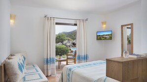 A bedroom with a bed, a wooden dresser, a television, and a view of a garden and mountains through the window. - Hotel with swimming pool near Baja Sardinia (Arzachena)
