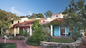 A row of Mediterranean-style buildings with stone walls and terracotta roofs, surrounded by lush gardens and a stone pathway. - Hotel with swimming pool near Baja Sardinia (Arzachena)