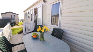 A covered patio with a round table, two chairs, a vase with flowers, and a bowl of fruit. - Beachcombers (Wareham)