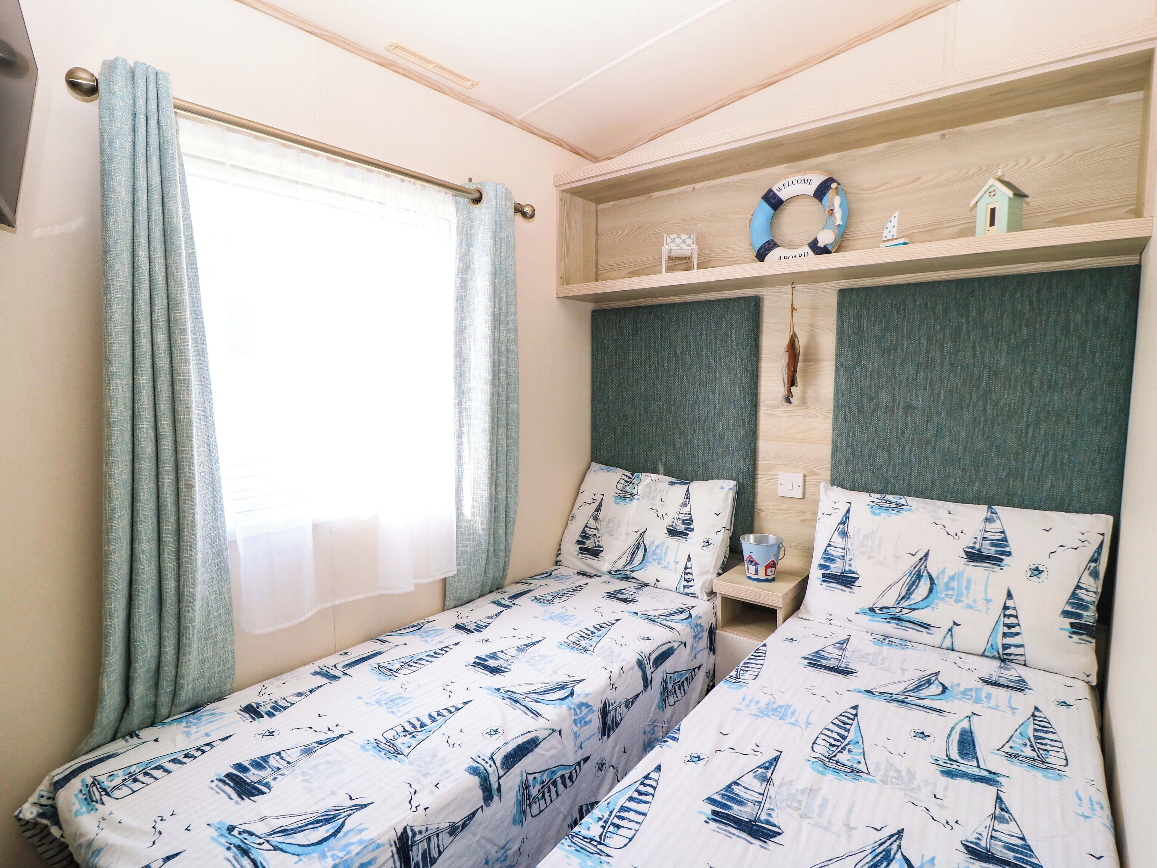 A bedroom with two beds, a window with curtains, a shelf with a welcome sign, and a small chair.