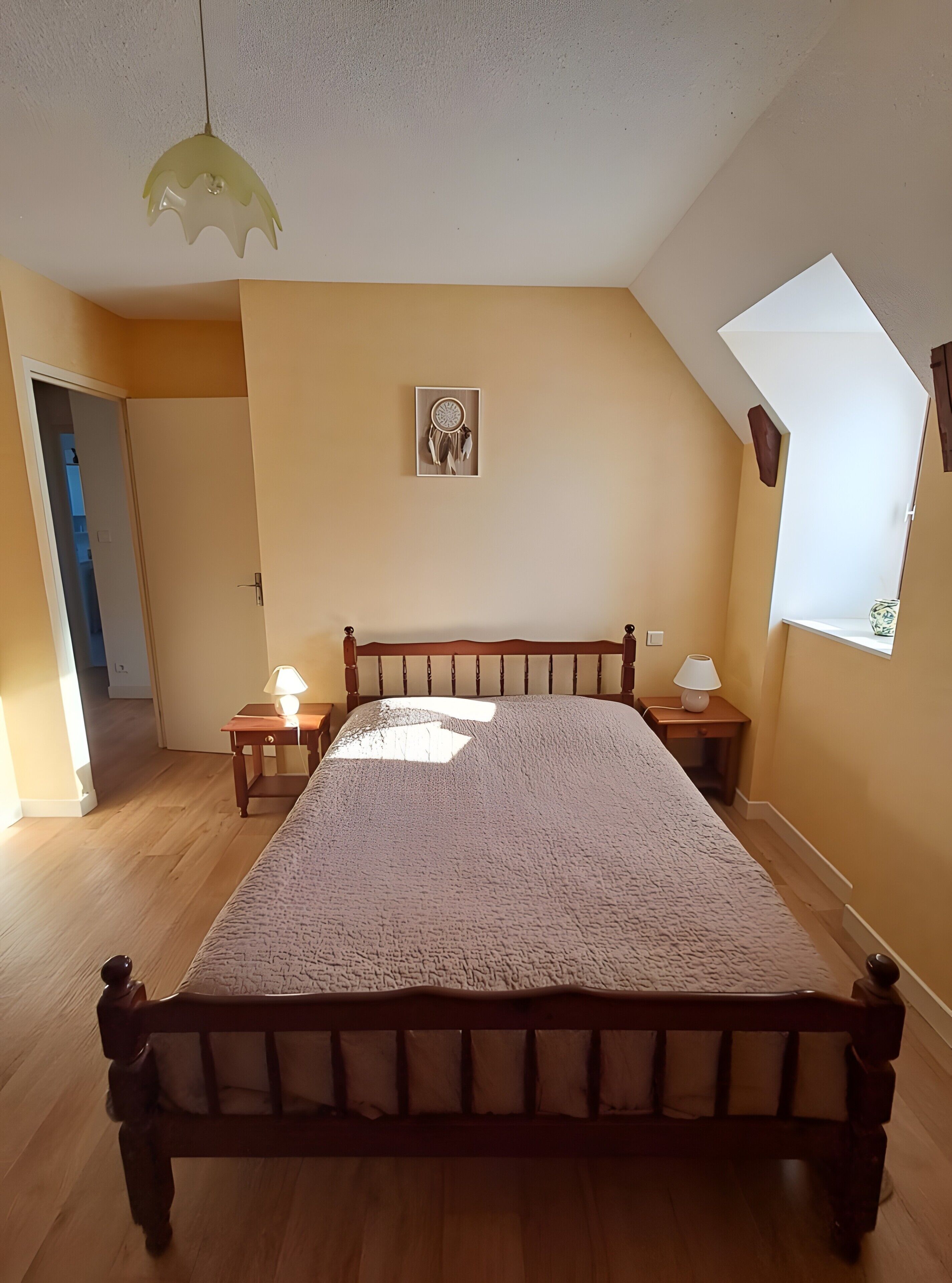 A bedroom with a wooden bed, two bedside tables with lamps, a dreamcatcher on the wall, and a sloped ceiling.