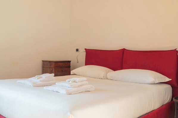 Un lit bien fait, avec du linge de lit blanc et des serviettes pliées, une table de chevet en bois et une tête de lit rouge.