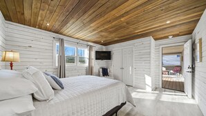 Une chambre avec un plafond en bois, un lit avec une literie blanche, un téléviseur fixé au mur et une porte donnant sur un espace extérieur.
