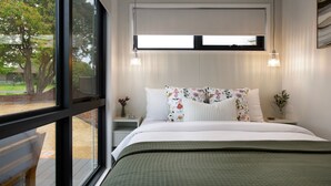 Une chambre à coucher moderne avec une grande fenêtre, un lit agrémenté d’oreillers à motifs floraux, une table de chevet et une petite table de nuit avec un vase.