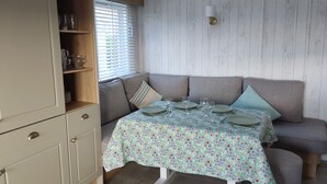 A dining area with a table set for four, a sofa, and a wooden cabinet with shelves. - Rockley Park - Harbour View, No 67 (Poole)