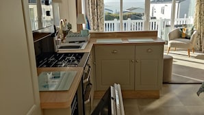 A kitchen with a gas stove, oven, and sink. There is a window with a view of houses and a balcony. - Rockley Park - Harbour View, No 67 (Poole)