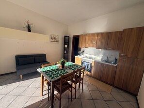 A compact kitchen with wooden cabinets, a dining table with a checkered tablecloth, and a sofa in a small living area. - L'Ulivo di Rollo-2 Terrazzi •Wi-Fi•Parcheggio (Capo Mimosa-rollo)