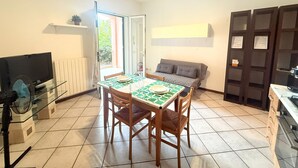 Uma sala de jantar com uma mesa posta para dois, um sofá, uma televisão e uma cozinha com fogão.