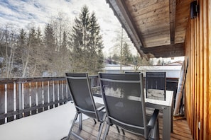 Een houten terras met twee stoelen en een tafel, met uitzicht op een besneeuwd landschap met bomen.