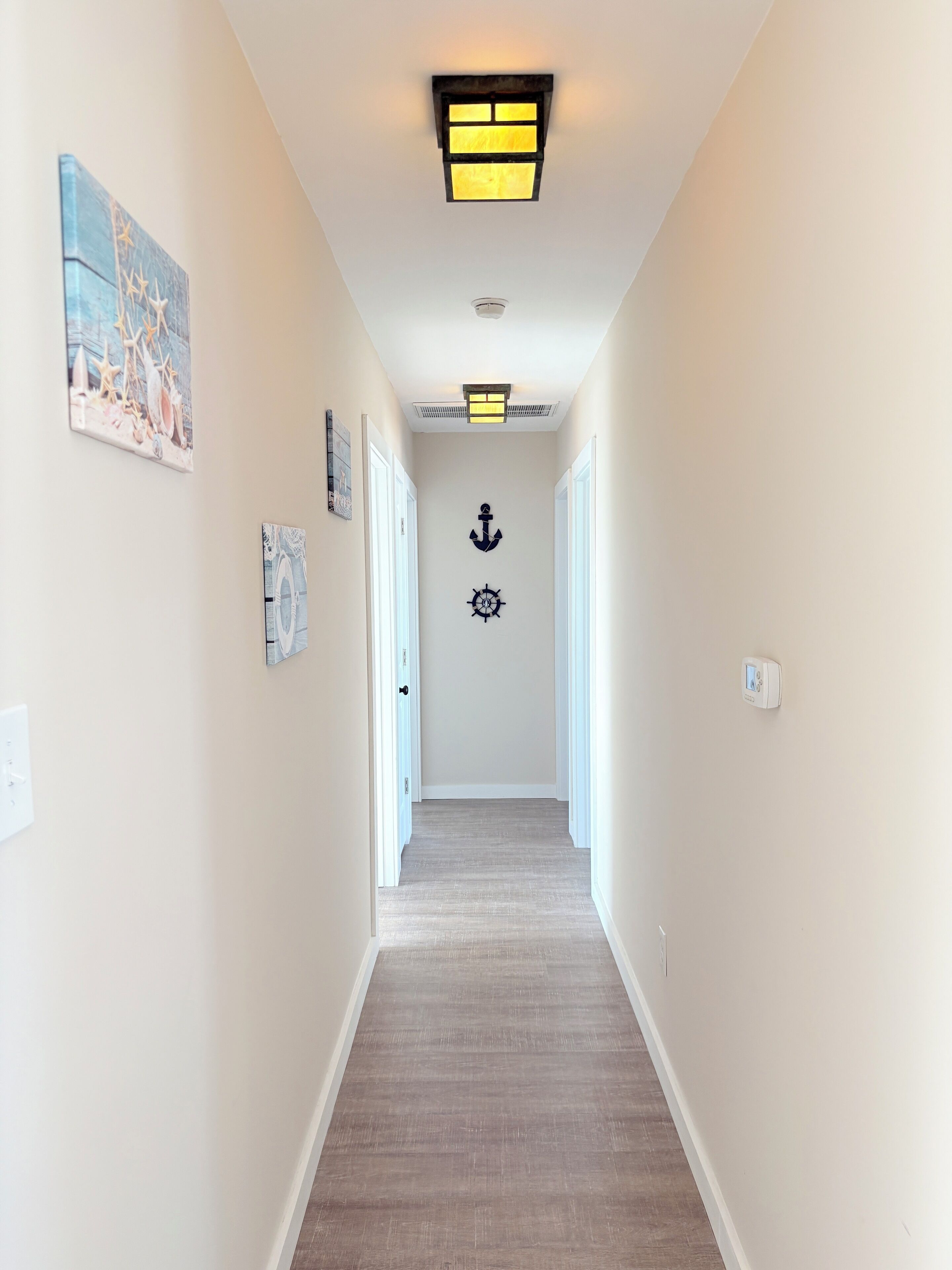 A hallway with a wooden floor, white walls, and a ceiling light. There are two doors on the left and a wall-mounted light fixture above the door.