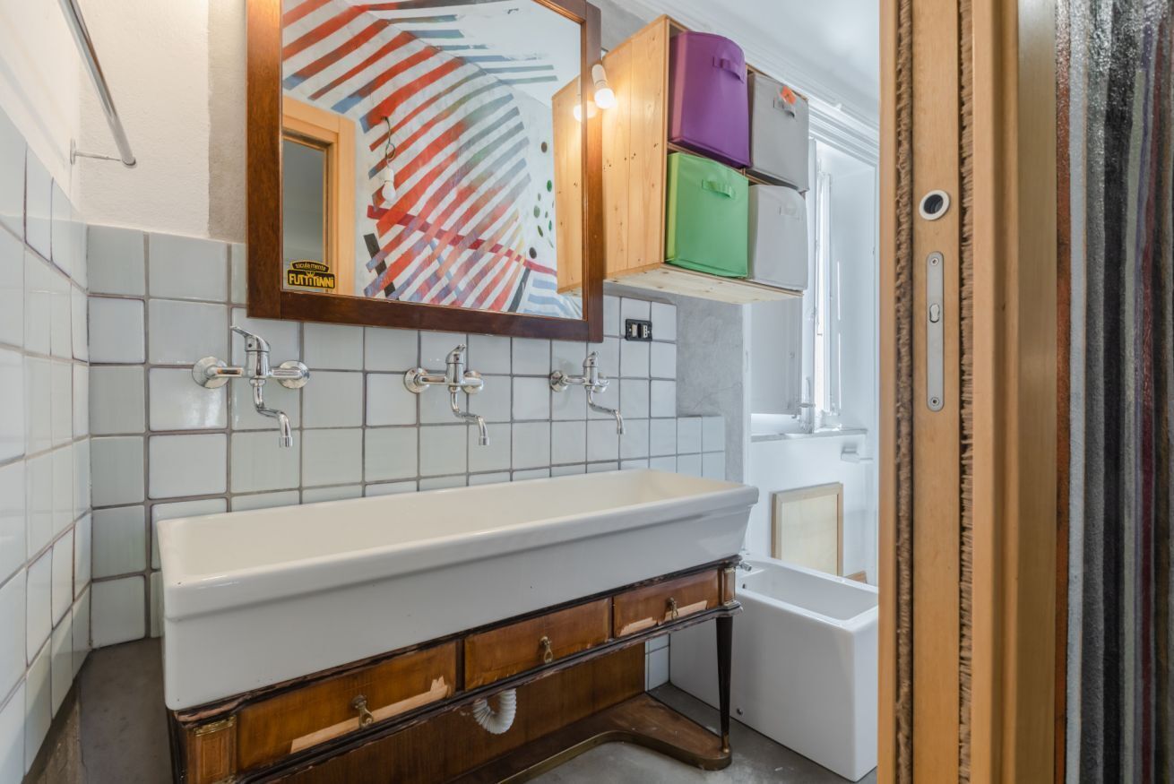 Une salle de bain avec un lavabo blanc, un meuble-lavabo en bois et un miroir orné d’un motif abstrait et coloré.