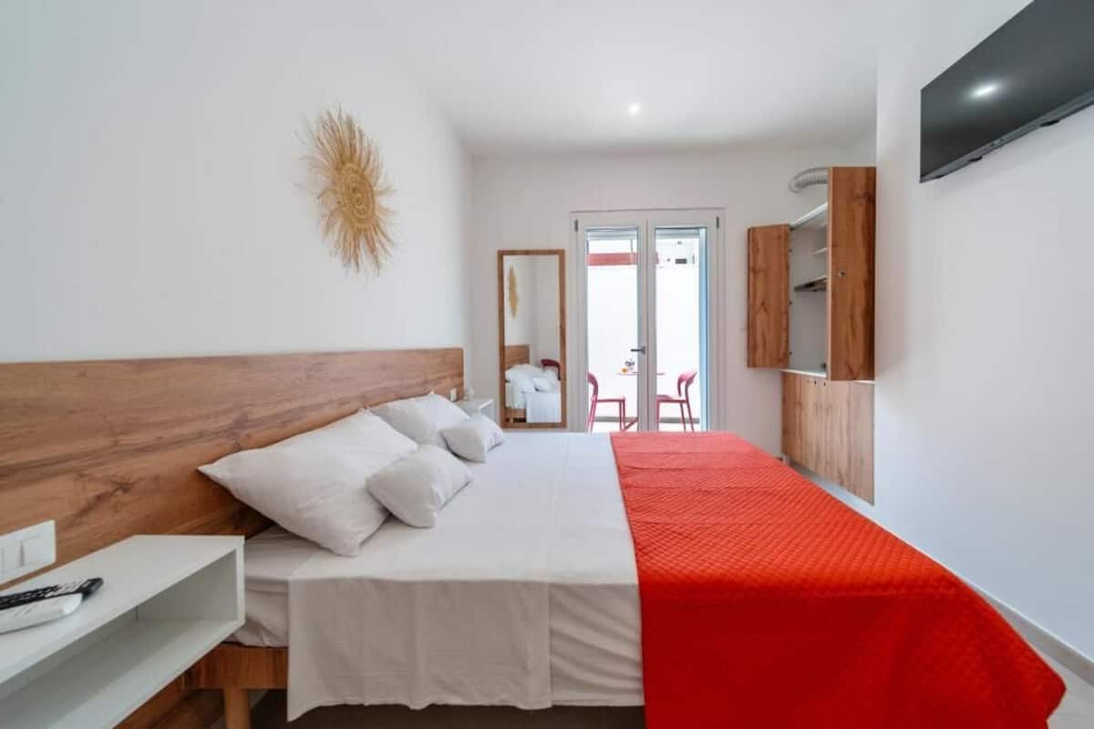 A bedroom with a wooden headboard, a bed with white bedding and a red blanket, a white bedside table, a mirror, and a TV mounted on the wall.