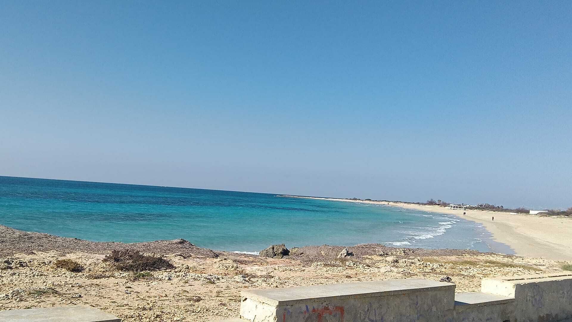Ein Strand mit klarem, blauem Wasser, sandigem Ufer und einer Betonbarriere im Vordergrund.