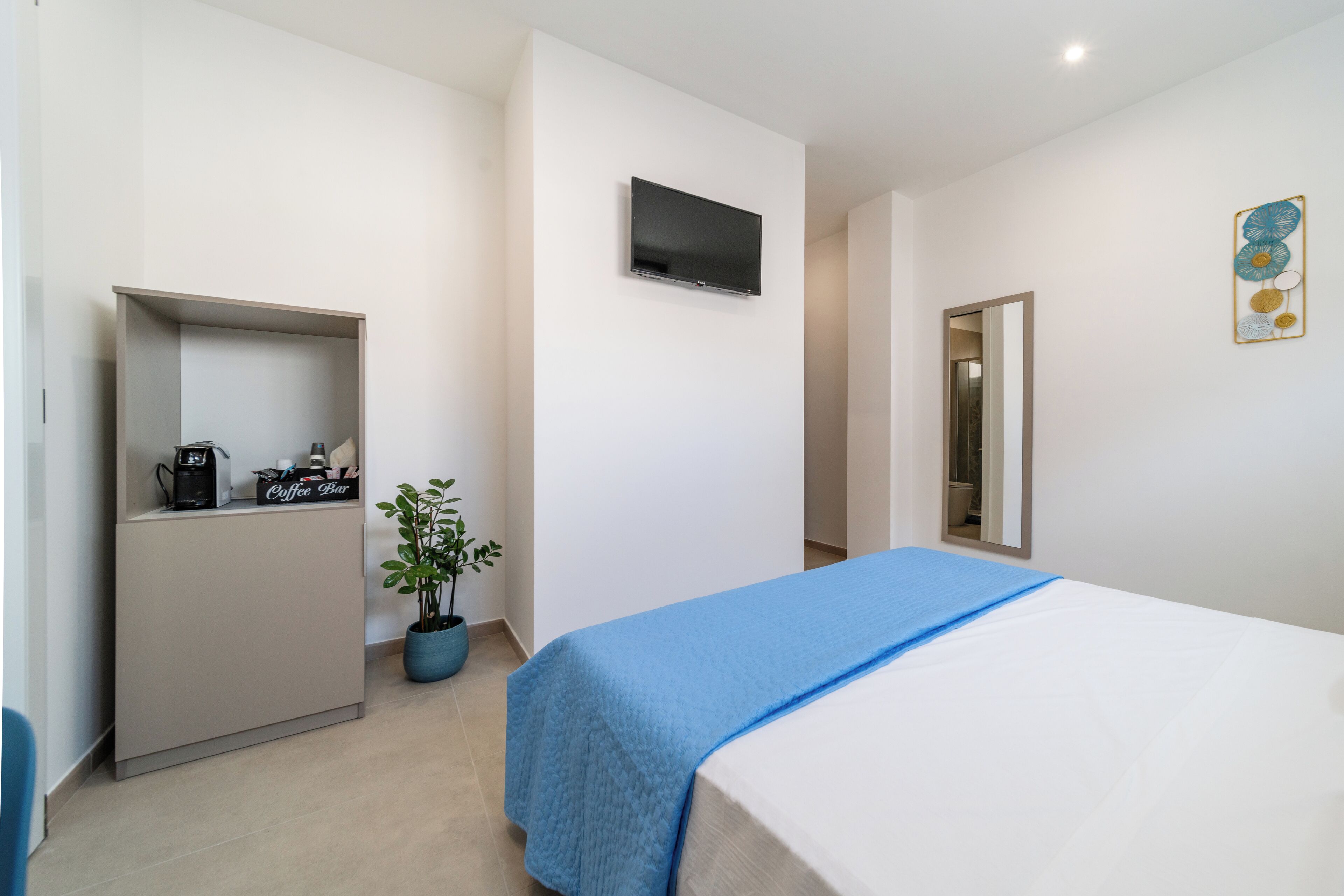 A hotel room with a bed, a coffee machine, a potted plant, and a flat-screen TV mounted on the wall.