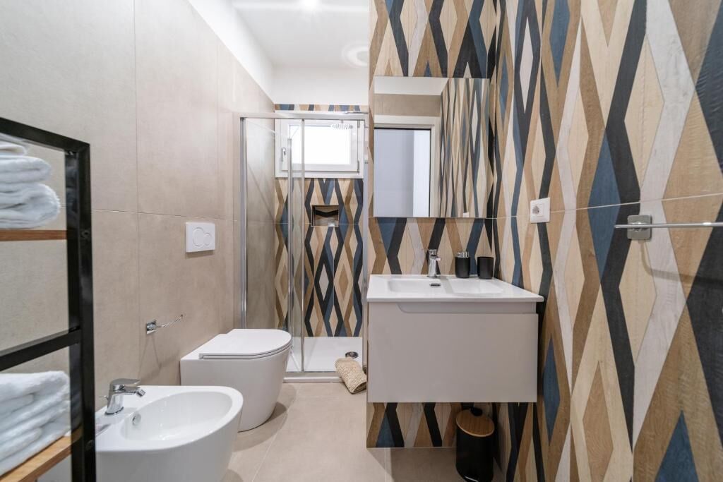 Un baño moderno con lavamanos, inodoro y bidé blancos. Presenta una pared con un diseño geométrico y un espejo sobre el lavamanos.