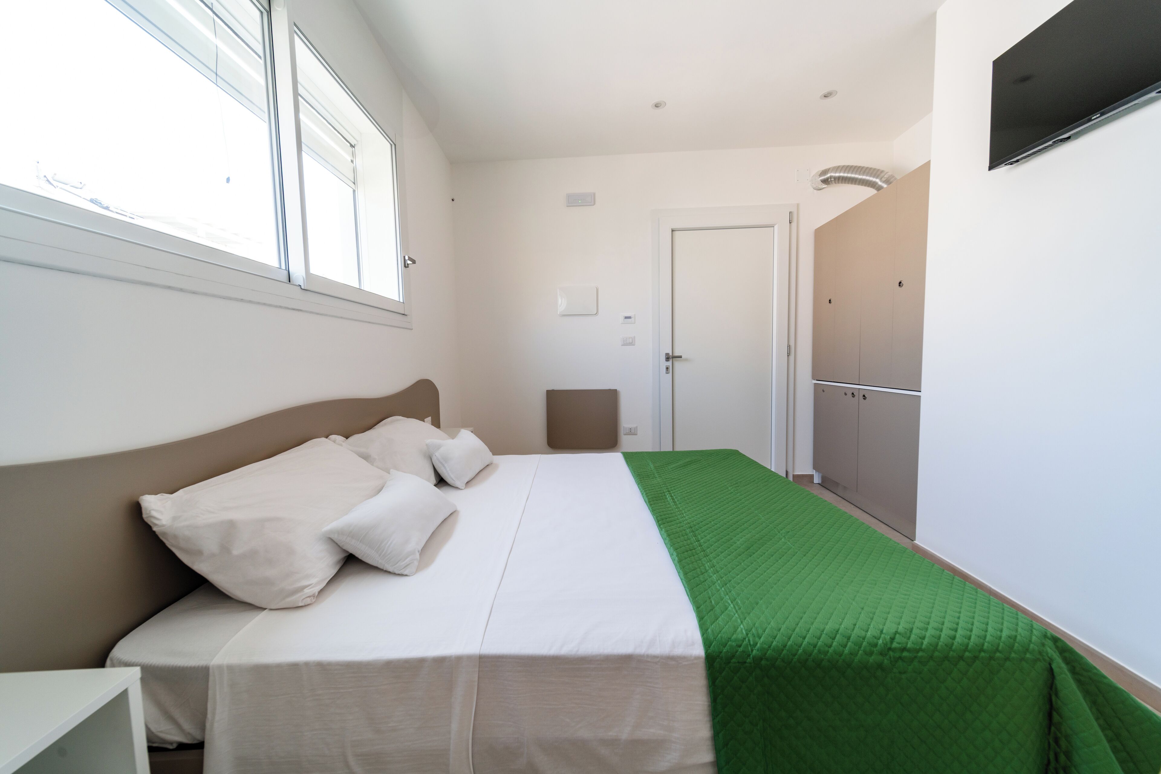 A bedroom with a bed, a green blanket, a white headboard, a nightstand, a wardrobe, and a door.
