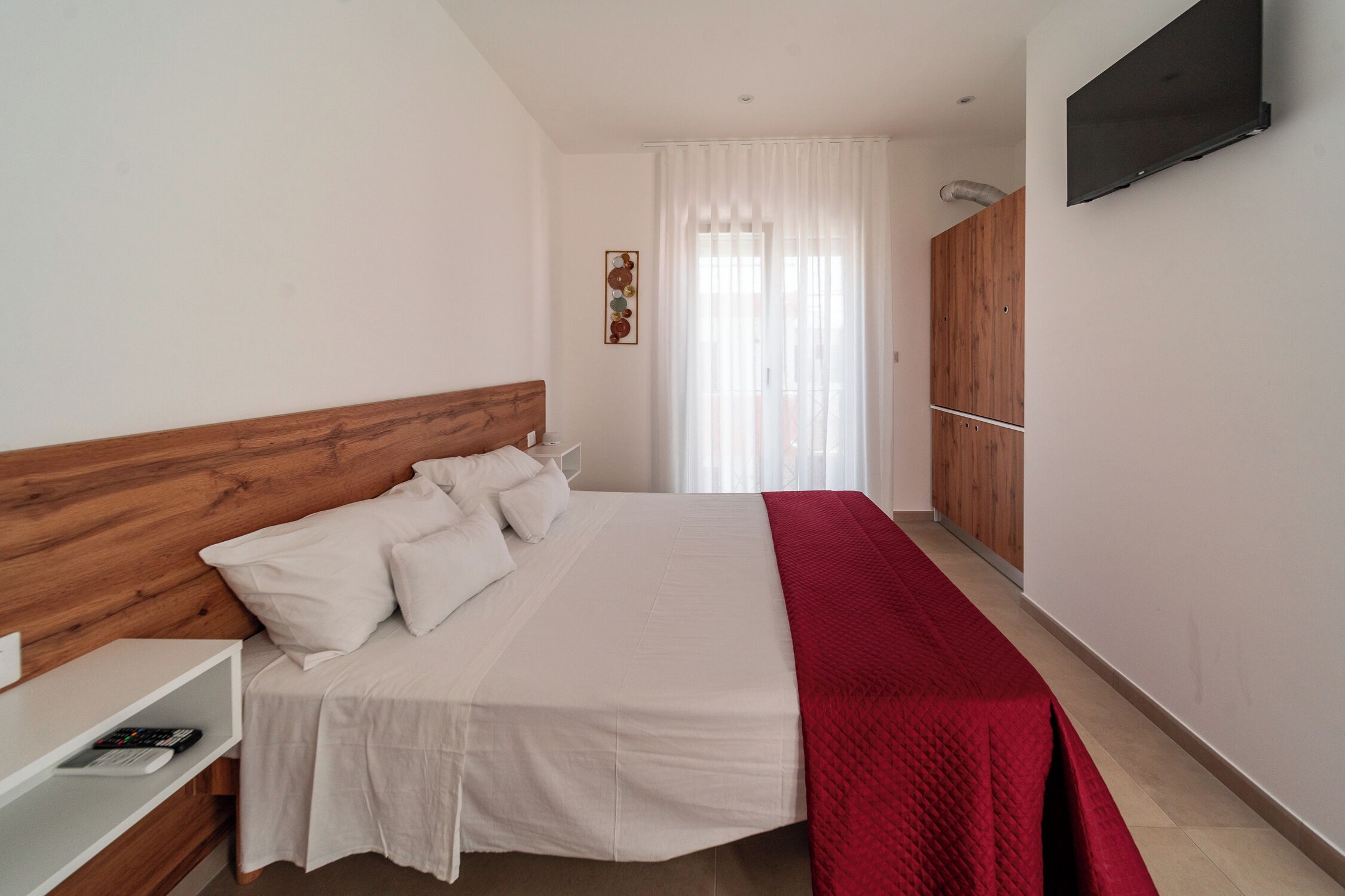 A bedroom with a wooden headboard, a white bedspread, and a red blanket. There is a TV mounted on the wall and a window with curtains.