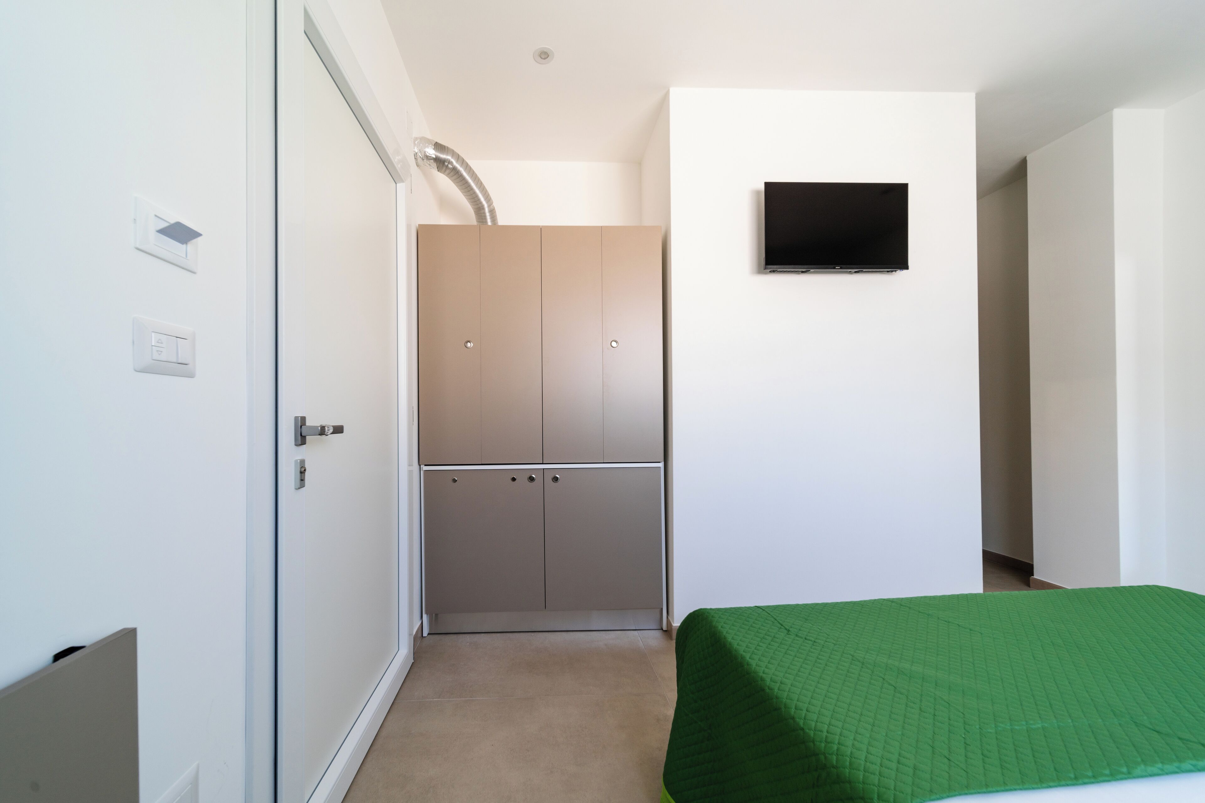 A small, modern room with a bed, a wardrobe, a television, and a ventilation duct.