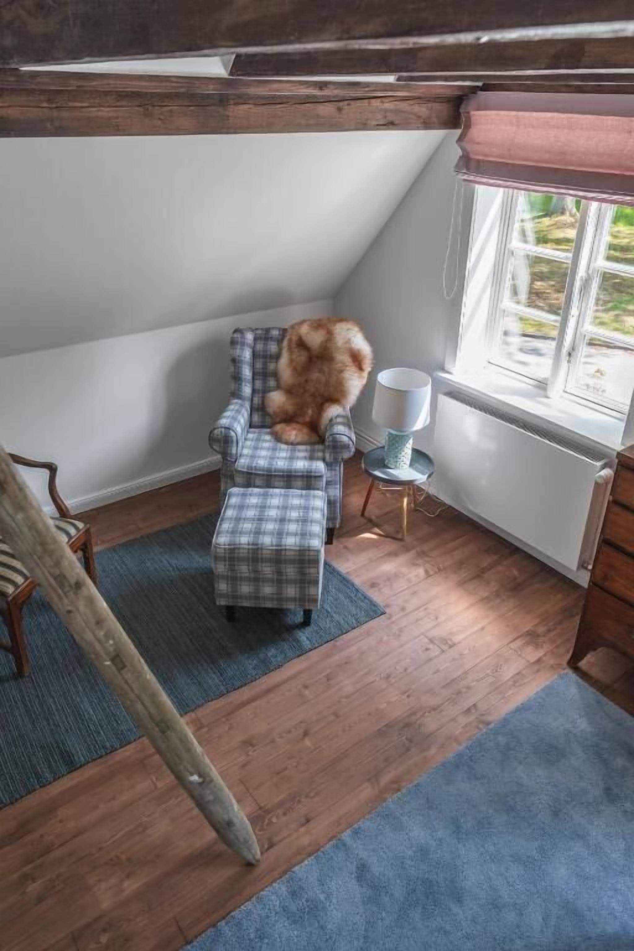Una habitación acogedora con un sillón y reposapiés de cuadros, una mesita con lámpara y una ventana con vistas a zonas verdes.