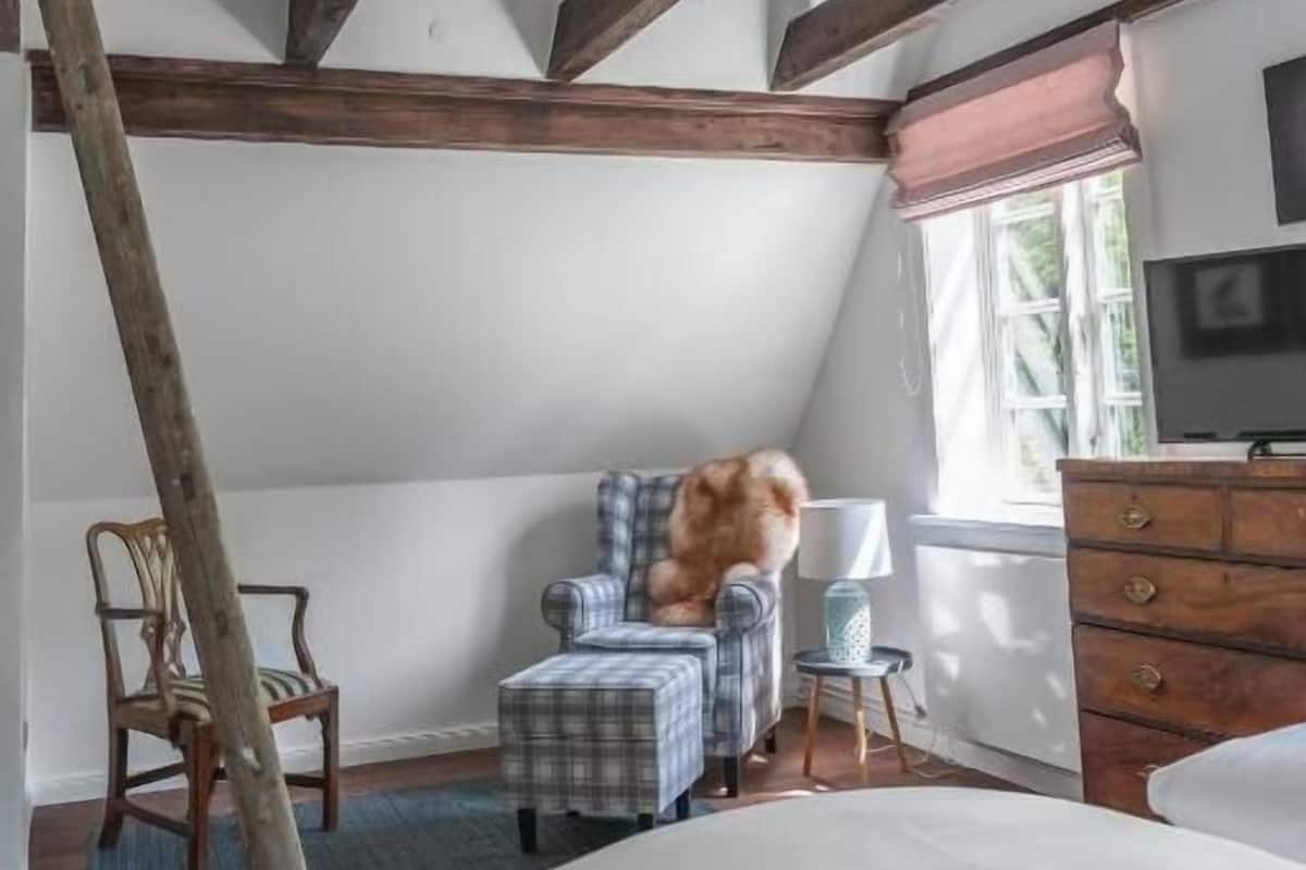A bedroom with a sloped ceiling, a bed, a television, a window with blinds, a chair, and a small table.