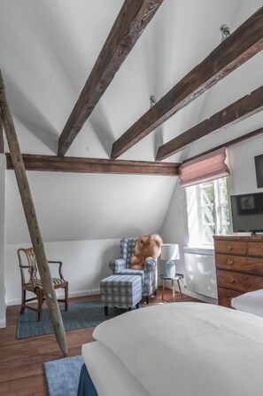 Une chambre avec un plafond en pente, un lit, une télévision, une fenêtre avec des stores, une chaise et une petite table.