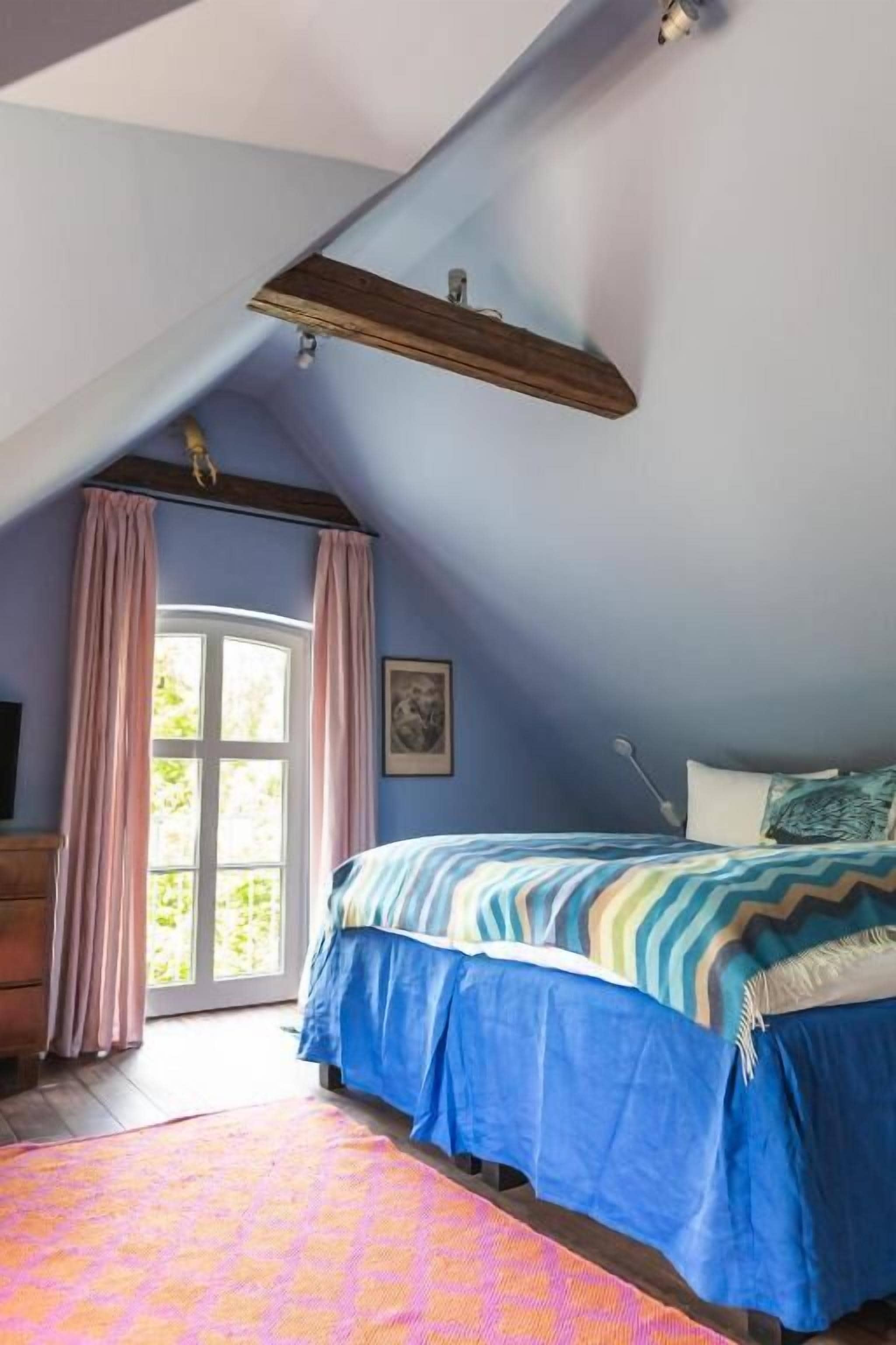 A bedroom with a sloped ceiling, a bed with a blue and white striped bedspread, a wooden headboard, and a window with pink curtains.