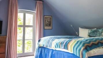 A bedroom with a sloped ceiling, a bed with a blue and white striped bedspread, a wooden headboard, and a window with pink curtains.