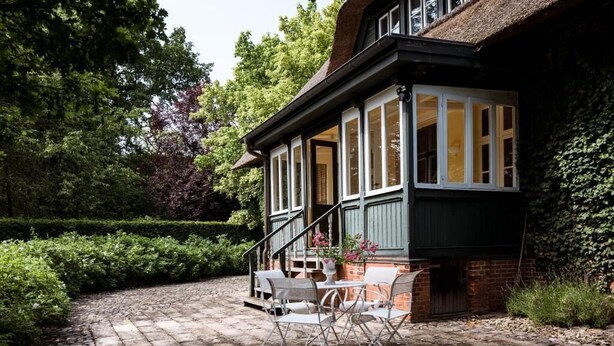 A covered patio with a brick floor, white chairs, and a table. - Alcor Exclusive Friedrichsruh (Aumuehle)