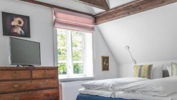 A bedroom with a sloped ceiling, wooden beams, a bed with white bedding, a dresser, a television, and a window with a view of greenery.