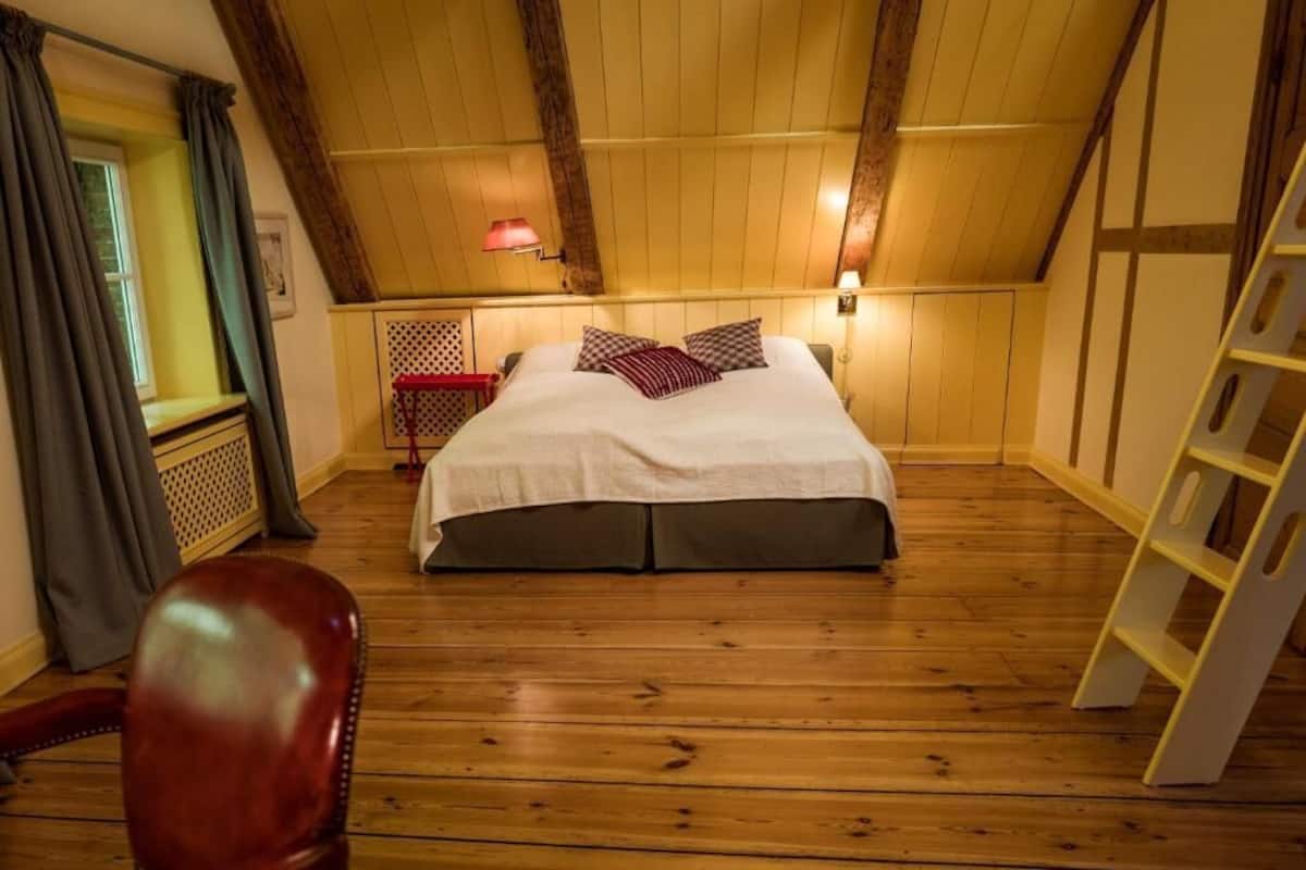 A bedroom with a wooden ceiling, a bed with a white bedspread and checkered pillows, a red chair, a ladder, and a window with curtains.