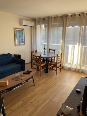A living room with a round wooden table, blue sofa, and a mounted air conditioner. - T2 air-conditioned 52 m² with parking - Near Thermes - Castle view - (Gréoux-les-Bains)