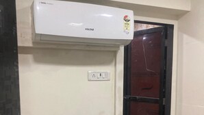 Ein Zimmer mit weißer Wand, einer weißen Klimaanlage, einer Tür mit schwarzem Rahmen und einer braunen Matte im Eingangsbereich.
